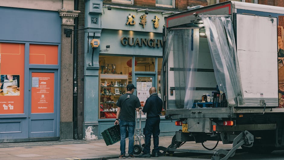 Two men wearing dark clothing are engaged in loading a moving truck parked outside a shop on a city street, with one man carrying a cardboard box and the other guiding with a hand trolley. The truck,