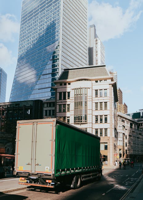 Two men wearing dark clothing are engaged in loading a moving truck parked outside a shop on a city street, with one man carrying a cardboard box and the other guiding with a hand trolley. The truck,