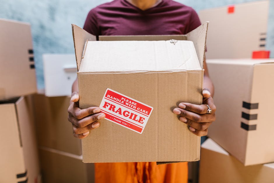 A person wearing a maroon top and orange trousers is holding an open cardboard box with both hands in a room filled with several similar boxes, some stacked and others standing singly. The boxes vary