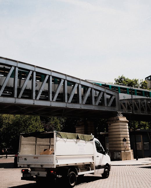 A white flatbed van with an open cargo area is parked on a paved street beneath a large elevated railway bridge structure made of metal with diagonal bracing, supported by two stone pillars. The cargo